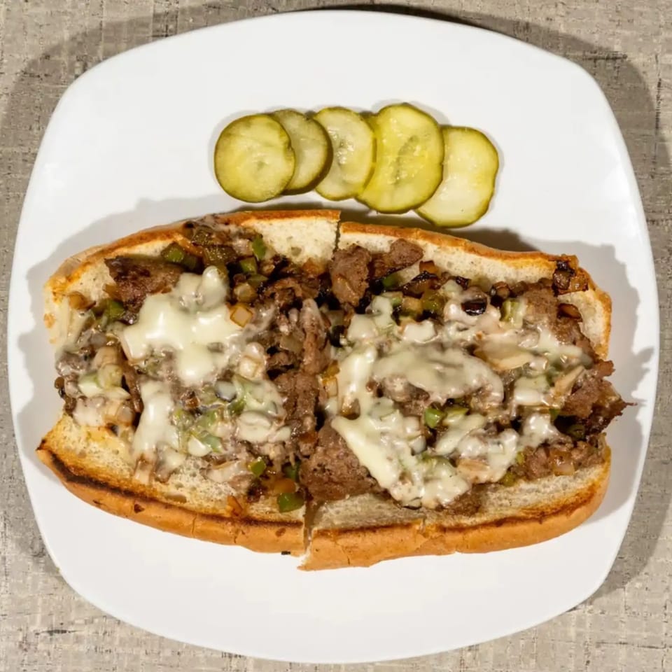 Cheesesteak sandwich with melted cheese, onions, and peppers, served with pickle slices on a white plate.