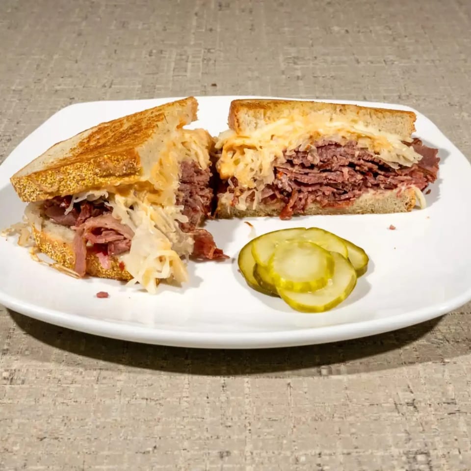 A grilled Reuben sandwich, cut in half, with pastrami, sauerkraut, and cheese, served with pickles.