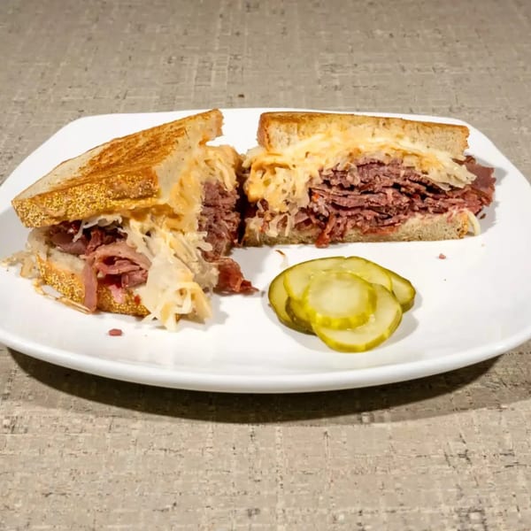 A Reuben sandwich cut in half, revealing corned beef, sauerkraut, and melted cheese, served with pickle slices.